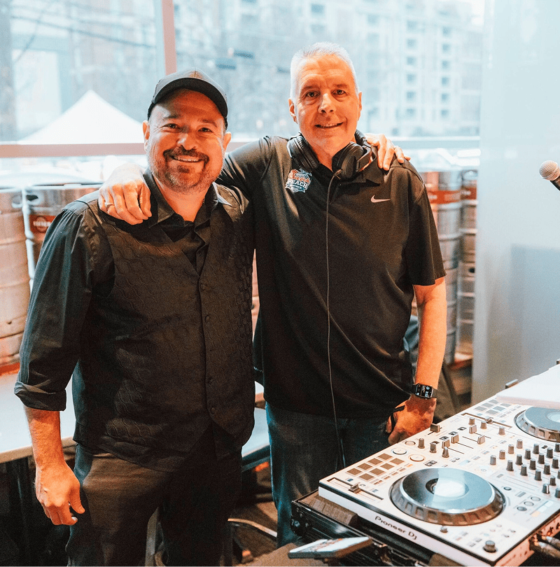 Two men smiling beside DJ equipment.
